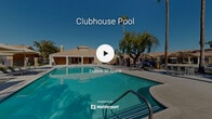 Located in Palm Desert, California, this outdoor community pool area offers a resort-style retreat beneath the sunny Coachella Valley skies. The centerpiece is a generously sized swimming pool with crystal-clear turquoise water, accessible via wide entry steps and handrail-equipped entry points, making it convenient for all users. Adjacent to the main pool is a separate spa, providing a dedicated space for relaxation. The expansive concrete pool deck is outfitted with multiple lounge chairs and shaded umbrella seating areas, offering ample space for sunbathing and socializing. A single-story clubhouse with a tile hip roof, columned covered patio, and warm stucco exterior sits directly alongside the pool, providing a welcoming indoor-outdoor gathering space. The entire area is enclosed by a black wrought-iron fence, ensuring privacy and security. Towering palm trees, mature shade trees, and vibrant desert-friendly landscaping with colorful flowering plants frame the perimeter, creating a lush, tropical ambiance. The surrounding backdrop of the San Jacinto Mountains is visible on the horizon, enhancing the scenic desert setting of this well-appointed community amenity.