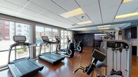 This fitness center is designed to support a complete workout experience within a spacious, single-floor layout. The space features polished concrete flooring in a rich reddish-brown tone, a dropped tile ceiling with recessed lighting, and bold purple accent walls that give the room a modern, energetic ambiance. Large floor-to-ceiling windows line one side of the facility, flooding the interior with natural light and offering open views of the surrounding neighborhood.

The cardio zone is well-stocked with multiple treadmills, elliptical trainers, and stationary bikes, all positioned to take advantage of the natural light from the large windows. The strength training area includes a range of Hammer Strength machines and cable systems, complemented by a full rack of dumbbells for free weight exercises. A dedicated stretching and functional training area provides ample open floor space for flexibility work and bodyweight exercises.

A built-in utility station with dark cabinetry, a granite countertop, and a sink offers a convenient area for equipment storage and personal needs. Mirrored doors lead to an adjacent room, extending the sense of space. The fitness center is well-organized, clean, and thoughtfully arranged to accommodate a variety of fitness routines in a comfortable, well-lit environment.