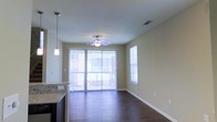 This three-floor property offers a well-designed living environment with thoughtful finishes throughout. The main living area showcases rich, dark hardwood flooring that adds warmth and contrast to the neutral, light beige walls. A ceiling fan with an integrated light fixture is centrally positioned, providing both airflow and ambient lighting to the space.

Natural light is a defining feature of this area, with a large sliding glass door fitted with vertical blinds allowing generous light to filter in while offering privacy control. An additional side window further enhances the brightness of the room. The sliding door likely provides access to an outdoor area or balcony, extending the functional living space.

A partial view of the kitchen reveals granite countertops and dark cabinetry, suggesting quality finishes consistent throughout the unit. A pendant light fixture hangs near the kitchen counter, adding a modern, stylish touch to the transitional space between the kitchen and living area.

Clean white baseboards frame the flooring neatly, and a ceiling vent indicates a central HVAC system. Spanning three floors, this property provides ample vertical space and separation between living zones.