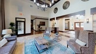 Located at 6900 Lake Woodlands Dr in Spring, TX, this sophisticated single-floor space serves as a welcoming community hub defined by its striking architectural details and refined interior design. The soaring ceilings are accented by dark exposed wood beams and complemented by an ornate wrought-iron chandelier, creating a grand yet inviting atmosphere throughout. Flooring transitions seamlessly between rich hardwood and natural stone tile, adding warmth and visual depth to the open layout. A dramatic stacked-stone accent wall serves as a stunning focal point, framed by lush greenery and decorative artwork that enhance the organic, upscale aesthetic. Floor-to-ceiling windows dressed with floor-length draping curtains flood the space with abundant natural light while offering expansive views of the surrounding landscaped outdoor areas and pool. The main lounge area is thoughtfully furnished with plush seating, layered rugs in teal and neutral tones, and carefully curated décor including circular medallion wall art and sculptural accents. A dedicated seating alcove provides a more intimate setting, ideal for conversations or waiting. A leasing or administrative office is visible to the side, equipped with workstations. The overall ambiance is elegant and modern, blending natural materials with contemporary furnishings for a polished, welcoming environment.