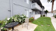 This ground-floor unit is part of a well-maintained multi-story residential building finished in a classic grey stucco exterior. The unit opens to a private outdoor patio space, paved with a concrete pad and furnished with a bistro-style table and chair — an ideal spot for relaxing or casual outdoor dining. The patio is framed by lush, manicured greenery, including a flourishing white rose bush and vibrant red flowering plants that add a welcoming burst of color to the exterior. A neatly trimmed grass lawn runs alongside the patio, enhancing the sense of a serene, well-kept outdoor environment. The unit is equipped with a wall-mounted air conditioning unit, ensuring comfort throughout the seasons. Large sliding windows allow natural light to filter into the interior. A white picket-style fence border adds a classic touch and defines the private outdoor area. Upper-floor units above feature white-railed balconies, contributing to the building's clean, cohesive aesthetic. Overall, the space combines practical residential amenities with pleasant outdoor living elements, making it a comfortable and inviting place to call home.