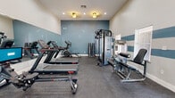 This fitness center offers a thoughtfully designed workout space that caters to a wide range of exercise needs. The room features soaring ceilings that lend an open, airy feel, while a full-length wall mirror along one side amplifies the sense of space and allows users to monitor their form during workouts. The walls are accented with a distinctive teal and white horizontal stripe design, adding a modern, energetic aesthetic to the space.

Natural light filters in through multiple large windows fitted with blinds, offering views of the surrounding greenery and keeping the room bright and inviting. Recessed lighting and warm ceiling-mounted fixtures provide ample illumination throughout. The flooring consists of durable rubber gym matting, ideal for both safety and equipment stability.

The cardio section is well-appointed with multiple Star Trac treadmills, an Echelon rowing machine, an Echelon stationary bike, and an elliptical trainer. For strength training, the space includes a TKO multi-functional cable machine, a TKO leg press/curl station, and a tiered dumbbell rack stocked with a range of free weights. A hand sanitizer station is conveniently positioned near the entrance, reflecting a commitment to cleanliness. The fitness center is accessible via a windowed door that connects to the outdoor area.