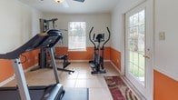 This dedicated fitness center is designed to support a comprehensive workout routine within a bright, well-organized space. The room features a distinctive two-tone color scheme, with bold orange on the lower half of the walls and crisp white above, creating an energetic atmosphere conducive to exercise. Light tile flooring runs throughout, providing a durable and easy-to-clean surface.

The space is outfitted with a notable selection of exercise equipment, including a treadmill, an elliptical trainer, a stationary cycle, and a Gold's Gym multi-station home gym system, offering users a full range of cardio and strength-training options. A large wall mirror on one side amplifies the sense of space and allows for form-checking during workouts.

Natural light enters generously through multiple windows and a glass door, which also provides an outdoor view of surrounding greenery. A ceiling fan overhead helps maintain air circulation during workouts. The glass door opens directly to the exterior, offering convenient access and ventilation. A decorative area rug near the entry adds a subtle finishing touch to this functional and well-appointed fitness space.