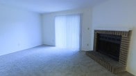 This single-floor residence offers a straightforward and functional layout designed for comfortable living. The main living area is a spacious, open room finished with wall-to-wall carpet that adds warmth and a soft underfoot feel throughout. A standout feature of this room is the classic brick fireplace, complete with a raised hearth, serving as a natural focal point and providing a cozy atmosphere. Adjacent to the fireplace, a large sliding glass door fitted with vertical blinds allows for generous natural light to flood the interior while also offering access to the outdoors. The clean white walls and simple architectural lines lend the space a bright and neutral canvas, making it easy to personalize. The overall ambiance is calm and understated, suited to a range of furnishing styles. The single-floor plan ensures easy accessibility and a cohesive flow throughout the home. With its combination of a brick fireplace, plush carpeting, and abundant natural light, this residence presents a welcoming and practical living environment.