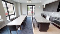 This contemporary single-floor space is thoughtfully designed to blend functionality with modern aesthetics. The open-plan layout seamlessly connects a spacious dining area with a well-appointed kitchen bar, creating an inviting environment for both casual and formal gatherings. The dining area features a long white rectangular table surrounded by sleek dark chairs, comfortably accommodating a large group. Overhead, a striking circular pendant light serves as a focal point, while recessed lighting throughout ensures the space is well-lit. Large black-framed windows line the walls, flooding the interior with natural light and offering views of the surrounding greenery and exterior building. The kitchen bar area is anchored by a generous granite-topped island with bar seating, complemented by dark cabinetry and a dedicated sink. Decorative pendant lights with a blue glass accent add a touch of artistic flair above the bar. The flooring transitions from patterned blue-grey carpet in the dining area to light hardwood near the kitchen, adding visual interest and defining each zone. The neutral grey walls and drop ceiling with acoustic tiles complete the clean, professional atmosphere of this versatile communal space.