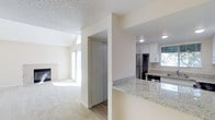 This single-floor space showcases an open-concept design that seamlessly connects the kitchen and living areas. The kitchen is well-appointed with granite stone countertops featuring subtle grey and white veining, white shaker-style upper cabinetry, and stainless steel appliances including a refrigerator and range. Natural light filters through a window above the kitchen sink, brightening the workspace. A peninsula counter separates the kitchen from the adjoining areas, offering additional prep and serving space.

A half-wall and structural column frame the transition between the kitchen and the rest of the layout, where wood-look flooring gives way to plush carpeting in the living area. The living space is anchored by a fireplace with a light-toned surround, adding a focal point to the room. Large windows allow generous natural light to fill the space. A closet with sliding doors is conveniently positioned near the living area. Throughout, the walls are finished in a neutral cream tone, and a textured ceiling runs consistently across the space, contributing to its cohesive, understated aesthetic.