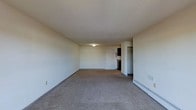 This single-floor space features a generously sized main living area finished with wall-to-wall carpeting in a neutral gray tone, providing a clean and understated foundation. The room is defined by smooth, white walls and a flat ceiling with a centrally mounted flush light fixture that provides overhead illumination. Along the far wall, a standard entry door is flanked by electrical outlets, keeping the space practical and functional. To the right side of the room, a baseboard heater runs along the base of the wall, offering an efficient heating solution. A floor vent is also visible nearby, suggesting a dual climate-control system. Toward the back of the space, an open closet area with a rod is partially visible, offering accessible storage. Adjacent to the closet, a darkened alcove hints at a connected room or kitchen area. The overall ambiance is simple and neutral, offering a blank canvas well-suited for personalization. The layout feels open and unobstructed, with ample floor space throughout the main living area. The minimalist design and light color palette contribute to a bright, airy feel despite the absence of windows in the immediate view.