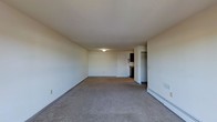 This single-floor space features a generously sized main living area finished with wall-to-wall carpeting in a neutral gray tone, providing a clean and understated foundation. The room is defined by smooth, white walls and a flat ceiling with a centrally mounted flush light fixture that provides overhead illumination. Along the far wall, a standard entry door is flanked by electrical outlets, keeping the space practical and functional. To the right side of the room, a baseboard heater runs along the base of the wall, offering an efficient heating solution. A floor vent is also visible nearby, suggesting a dual climate-control system. Toward the back of the space, an open closet area with a rod is partially visible, offering accessible storage. Adjacent to the closet, a darkened alcove hints at a connected room or kitchen area. The overall ambiance is simple and neutral, offering a blank canvas well-suited for personalization. The layout feels open and unobstructed, with ample floor space throughout the main living area. The minimalist design and light color palette contribute to a bright, airy feel despite the absence of windows in the immediate view.