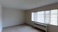 This single-floor space presents a clean, understated interior defined by its simple and functional design. The main room is finished with light-colored carpeting and white walls, creating a neutral, versatile backdrop. A textured popcorn ceiling adds subtle character overhead while maintaining the room's airy feel. A large picture window dominates one wall, fitted with vertical blinds that allow for adjustable light control. The window offers a view of an outdoor fenced area with greenery visible beyond, bringing a connection to the exterior environment. Beneath the window sits a baseboard radiator heating unit, providing an efficient heat source positioned to maximize warmth distribution throughout the room. The overall layout is open and uncluttered, offering flexibility in how the space can be arranged and used. The combination of natural light from the oversized window and the bright white walls contributes to a spacious and well-lit atmosphere. The space presents a straightforward, move-in-ready environment suited to a variety of uses.