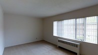 This single-floor space presents a clean, understated interior defined by its simple and functional design. The main room is finished with light-colored carpeting and white walls, creating a neutral, versatile backdrop. A textured popcorn ceiling adds subtle character overhead while maintaining the room's airy feel. A large picture window dominates one wall, fitted with vertical blinds that allow for adjustable light control. The window offers a view of an outdoor fenced area with greenery visible beyond, bringing a connection to the exterior environment. Beneath the window sits a baseboard radiator heating unit, providing an efficient heat source positioned to maximize warmth distribution throughout the room. The overall layout is open and uncluttered, offering flexibility in how the space can be arranged and used. The combination of natural light from the oversized window and the bright white walls contributes to a spacious and well-lit atmosphere. The space presents a straightforward, move-in-ready environment suited to a variety of uses.