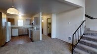 This two-floor residential space offers a practical and straightforward layout designed for comfortable everyday living. The main level features an open-plan design where the kitchen flows directly into the living area, creating a cohesive and accessible common space. The kitchen is equipped with light wood cabinetry, a double sink positioned near a window that brings in natural light, an electric range, and a central island or peninsula that provides additional counter space and separates the kitchen from the living area. The flooring in the kitchen is hard surface, transitioning to plush carpeting throughout the living area, adding warmth and comfort underfoot. A wall-mounted heating unit is conveniently located in the living space. A doorway visible from the main area leads to what appears to be a utility or laundry room, adding functional storage and convenience. The staircase, accented with a black metal railing, provides access to the second floor. The overall ambiance of the main level is clean, neutral, and move-in ready, with white walls and a simple, uncluttered aesthetic that offers a blank canvas for personalization.