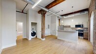 Located at 237-243 Furnace St in Akron, Ohio, this two-bedroom, one-bathroom loft unit masterfully combines historic industrial character with contemporary updates. The open-concept living and kitchen area features light hardwood flooring, soaring ceilings with exposed metal ductwork, and original heavy timber columns that speak to the building's storied past. The kitchen is well-appointed with stainless steel appliances—including a refrigerator, electric range, built-in microwave, and dishwasher—complemented by white cabinetry, a subway tile backsplash, and a peninsula with a white countertop that defines the space. The primary bedroom showcases striking exposed brick walls with arched alcoves, adding dramatic architectural texture, while plush carpeting provides comfort underfoot. The second bedroom retains the loft's industrial character through original timber posts framing the carpeted room. The bathroom offers a clean, modern aesthetic with a tiled tub-and-shower combination, a wood-toned vanity, and light flooring. Throughout the unit, the interplay of raw brick, aged wood beams, and crisp white walls creates a warm yet distinctly urban ambiance that reflects the character of this converted industrial building.
