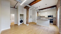 Located at 237-243 Furnace St in Akron, Ohio, this two-bedroom, one-bathroom loft unit masterfully combines historic industrial character with contemporary updates. The open-concept living and kitchen area features light hardwood flooring, soaring ceilings with exposed metal ductwork, and original heavy timber columns that speak to the building's storied past. The kitchen is well-appointed with stainless steel appliances—including a refrigerator, electric range, built-in microwave, and dishwasher—complemented by white cabinetry, a subway tile backsplash, and a peninsula with a white countertop that defines the space. The primary bedroom showcases striking exposed brick walls with arched alcoves, adding dramatic architectural texture, while plush carpeting provides comfort underfoot. The second bedroom retains the loft's industrial character through original timber posts framing the carpeted room. The bathroom offers a clean, modern aesthetic with a tiled tub-and-shower combination, a wood-toned vanity, and light flooring. Throughout the unit, the interplay of raw brick, aged wood beams, and crisp white walls creates a warm yet distinctly urban ambiance that reflects the character of this converted industrial building.
