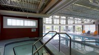 This single-level indoor aquatic facility offers a well-appointed environment designed for both recreation and relaxation. At the heart of the space is a generously sized indoor swimming pool, complemented by a dedicated spa with clearly posted capacity guidelines, ensuring a safe and comfortable experience for all users. The pool area is framed by an impressive structural ceiling featuring exposed steel beams and trusses, lending an industrial yet polished aesthetic to the facility. Floor-to-ceiling windows line the perimeter, inviting abundant natural light and providing views of the surrounding greenery, creating a bright and airy atmosphere even while indoors. The spa is conveniently accessible via stainless steel handrails, reflecting a strong focus on accessibility and safety throughout. A deep red accent wall adds a bold architectural contrast to the otherwise neutral tones of the space. Beyond the pool, a seating area near the windows offers a relaxed spot to rest and take in the outdoor scenery. The combination of high ceilings, generous glazing, and thoughtfully arranged amenities makes this aquatic space both functional and inviting for swimmers and spa-goers alike.