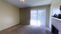 This single-floor space features a living area finished with wall-to-wall carpeting that adds warmth underfoot. A large sliding glass door dominates one wall, dressed with full-length vertical blinds that allow for adjustable light control while providing access to an outdoor area. Natural light filters through the blinds, brightening the room. The walls are painted in a neutral tan tone, complementing the neutral carpet. A ceiling fan with an integrated light fixture is centrally mounted, offering both airflow and ambient lighting. The textured ceiling adds subtle visual interest overhead. Electrical outlets and a light switch are positioned along the walls. The overall ambiance is understated and functional, offering a blank canvas well-suited for a variety of furnishing arrangements.
