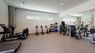 This fitness center is designed to support a full range of workout routines within a clean, modern environment. The space is anchored by a dramatic floor-to-ceiling window wall that floods the interior with natural light and offers views of the surrounding outdoor greenery, creating an energizing atmosphere for exercise. The high ceilings amplify the sense of openness throughout.

The cardio zone is positioned along the window frontage and features multiple treadmills, a stair climber, and stationary cycling bikes, each equipped with personal display screens. A dedicated strength training area showcases a multi-station cable and weight machine, a squat/power rack with a flat bench, and additional resistance equipment — all reflected in a full wall of mirrors that visually expands the space and aids in form correction during workouts.

A free weights section includes a well-organized rack stocked with kettlebells and medicine balls in various sizes. The flooring throughout is durable rubber-backed carpet, appropriate for both cardio and strength activities. Recessed lighting complements the natural light, ensuring the space remains bright at all hours. A small ancillary alcove is also present, complete with a sink, providing a convenient area for hygiene or towel service. Overall, this fitness center is thoughtfully appointed and well-suited for comprehensive daily training.