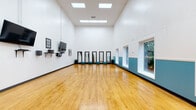 This single-floor studio is thoughtfully designed to support movement-based activities, offering a generous open floor plan with gleaming hardwood floors that reflect both natural and artificial light. The soaring ceilings create an airy, expansive atmosphere, making the space feel even larger and more welcoming for a variety of uses.

The crisp white walls are accented by a teal lower wainscoting detail along one side, adding a touch of color and visual interest to the otherwise clean, minimalist interior. A row of tall, black-framed full-length mirrors lines one wall, accompanied by small teal cushioned stools at their bases — ideal for stretching or brief rest between sessions. Lush green potted plants flank the mirror arrangement, introducing a refreshing natural element.

Recessed panel lighting in the ceiling ensures even, bright illumination throughout, while multiple large windows along the side walls invite ample natural light and offer views of the surrounding greenery. Wall-mounted flat-screen televisions and audio-visual equipment on bracket shelves provide the technological infrastructure needed for instruction or media playback. An open metal shelving unit and a dedicated door to an adjacent area add functional storage and accessibility to the layout. The overall ambiance is clean, professional, and energizing.