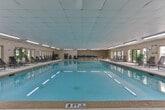 Located in Decatur, Georgia, this single-floor indoor pool facility at 4946 Snapfinger Woods Dr offers a well-appointed aquatic environment designed for both recreation and relaxation. The centerpiece of the space is a generously sized indoor swimming pool with clear, turquoise water, marked with lane indicators and outfitted with entry handrails for convenient access. The pool area is enclosed within a bright, elongated room featuring a suspended tile ceiling equipped with rows of fluorescent lighting that evenly illuminate the space.

A wide, textured pool deck runs the length of the facility, furnished with multiple reclining lounge chairs positioned along the windowed wall. A series of large windows lines one side of the room, allowing natural light to filter in and offering views of the surrounding greenery. Decorative potted tropical plants, including a prominent palm, add a welcoming, resort-like ambiance to the interior.

Access to an outdoor patio area is provided through double glass doors, where additional seating and a secondary outdoor pool area can be seen. The facility also features posted pool rules and safety equipment, including a life ring. The overall design prioritizes both functionality and comfort, making it a practical and inviting aquatic amenity.