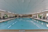 Located in Decatur, Georgia, this single-floor indoor pool facility at 4946 Snapfinger Woods Dr offers a well-appointed aquatic environment designed for both recreation and relaxation. The centerpiece of the space is a generously sized indoor swimming pool with clear, turquoise water, marked with lane indicators and outfitted with entry handrails for convenient access. The pool area is enclosed within a bright, elongated room featuring a suspended tile ceiling equipped with rows of fluorescent lighting that evenly illuminate the space.

A wide, textured pool deck runs the length of the facility, furnished with multiple reclining lounge chairs positioned along the windowed wall. A series of large windows lines one side of the room, allowing natural light to filter in and offering views of the surrounding greenery. Decorative potted tropical plants, including a prominent palm, add a welcoming, resort-like ambiance to the interior.

Access to an outdoor patio area is provided through double glass doors, where additional seating and a secondary outdoor pool area can be seen. The facility also features posted pool rules and safety equipment, including a life ring. The overall design prioritizes both functionality and comfort, making it a practical and inviting aquatic amenity.