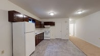 This single-floor unit offers a functional and straightforward layout, with distinct areas for living, cooking, sleeping, and bathing. The kitchen is equipped with dark brown cabinetry spanning both upper and lower sections, providing ample storage space. White appliances, including a refrigerator, electric range, and an under-cabinet range hood, complement the light countertops and stone-look vinyl tile flooring. A window above the sink allows for a modest amount of natural light to enter the space.

The kitchen opens directly into a combined living area, where the flooring transitions from vinyl tile to neutral-toned carpet. This open-concept arrangement creates a generous, flexible space suited for both lounging and dining. Baseboard heating runs along the walls, and a wall-mounted air conditioning unit provides additional climate control. Flush-mount ceiling lighting keeps the area well lit.

The bedroom is a comfortable, carpeted room featuring a ceiling fan with an integrated light fixture, a baseboard heater, and a window that brings in natural light. A panel box is conveniently located on the wall near the entry door.

The bathroom is finished with light ceramic tile flooring and full white tile surround in the tub/shower combo. A small window above the tub provides ventilation and additional light, completing this clean, no-frills space.