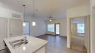 This single-story space showcases an open-concept layout that seamlessly connects the kitchen and living areas. The kitchen features a granite-style speckled countertop with a stainless steel sink and is illuminated by two modern pendant lights, creating a functional yet stylish workspace. White cabinetry adds a clean, bright aesthetic to the area.

The living space extends naturally from the kitchen, finished with wood-look plank flooring that adds warmth throughout. A ceiling fan is centrally positioned overhead, offering both practical airflow and a subtle design element. A glass-paneled door at the far end of the living area allows natural light to filter in, enhancing the open and airy feel of the space.

Neutral white walls throughout provide a versatile backdrop, while doorways visible on both sides of the living area lead to additional rooms, suggesting a well-organized floor plan. One adjoining room features carpeted flooring, offering a softer surface option within the unit. The overall ambiance is clean, modern, and move-in ready, with consistent finishes that create a cohesive living environment.