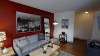 This single-floor space makes a strong visual impression with its deep crimson accent wall, which serves as a dramatic backdrop in the main living area. The living room is anchored by a comfortable grey tufted sofa adorned with casual throw pillows, paired with a wooden coffee table that adds a warm, grounded feel. A full-length mirror in a sleek black frame reflects light and adds depth to the room. Overhead, an arc floor lamp provides warm, focused lighting that complements the space's cozy ambiance. The flooring throughout features rich, dark hardwood planks that add warmth and continuity. A Parisian theme runs through the décor, with large black-and-white photographs and prints of the Eiffel Tower and the Paris skyline displayed prominently on the walls, lending a cosmopolitan and cultured character to the interior. A compact side table with open shelving provides functional storage, while a leopard-print bean bag chair introduces a playful, eclectic touch. The textured ceiling adds subtle architectural interest. Overall, the space balances bold design choices with practical furnishings, creating an inviting and personality-rich environment.