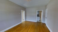 This single-floor space offers a straightforward and functional layout designed for comfortable everyday living. The main living area is characterized by warm-toned hardwood floors that run throughout, adding a classic and timeless quality to the interior. The walls are finished in a neutral gray tone, complemented by crisp white baseboards and door trim that provide clean visual contrast and a well-maintained appearance.

The living area is notably open, with generous ceiling height that contributes to an airy feel. Two doorways lead from the main room to adjacent spaces — one opening to what appears to be an additional room or hallway, and the other providing direct access to the kitchen. The kitchen, visible through the doorway, features white cabinetry and appliances, suggesting a practical and efficient layout.

The overall design of the space is simple and uncluttered, making it highly adaptable for a variety of uses and furnishing arrangements. Natural light appears to reach the interior, enhancing the warmth of the hardwood flooring and brightening the neutral palette. The combination of classic hardwood floors, neutral tones, and white trim creates a cohesive and inviting atmosphere throughout the space.