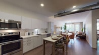 This single-floor residence showcases a thoughtfully designed open-concept layout that seamlessly connects the kitchen, dining, and living areas. The kitchen is outfitted with sleek flat-front white cabinetry, stainless steel appliances including a range, built-in microwave, and dishwasher, complemented by clean white countertops and a subway tile backsplash. An exposed cylindrical ventilation duct adds an industrial accent to the otherwise minimalist aesthetic. A central kitchen island with bar seating bridges the cooking and dining spaces. The open living area is bright and welcoming, benefiting from large windows that invite generous natural light and offer views of lush outdoor greenery. Wide-plank hardwood-style flooring flows consistently throughout the space, enhancing the sense of continuity. The bedroom is generously proportioned, featuring a dedicated workspace area, a large window with natural light, and ample room for both rest and productivity. The bathroom presents a clean, contemporary design with a white vanity, a large mirror, pendant lighting, and neutral grey wall tones. Recessed lighting throughout the residence ensures a well-lit, modern ambiance across all areas.