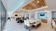 This single-floor space showcases a thoughtfully designed open-concept layout that seamlessly integrates multiple functional zones into one cohesive environment. The design immediately draws the eye upward to a striking coffered ceiling clad in warm natural wood planking, accented by embedded LED strip lighting that casts a soft, ambient glow throughout the room.

The dining area serves as a central focal point, anchored by a large rectangular white table surrounded by cream upholstered chairs with wood-toned legs. Above it, a dramatic cluster of oversized glass globe pendant lights adds an elegant, modern touch. Adjacent to the dining space is a wet bar or kitchenette area, finished with light blue tile backsplash, stainless steel appliances, and a wall-mounted display screen.

Flowing into the background, a game room zone features a regulation-style billiards table set against a decorative mosaic tile accent wall with its own mounted television. A casual lounge area with contemporary seating rounds out the space. Light-toned large-format tile flooring unifies all zones, while recessed lighting and pendant fixtures maintain a bright, welcoming atmosphere throughout.
