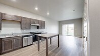 Located at 522 Sweetgrass Dr in Brookings, SD, this one-bedroom, one-bathroom unit offers a thoughtfully designed single-floor layout that maximizes functionality and comfort. The open-concept kitchen and living area serves as the heart of the space, featuring light wood-tone flooring throughout. The kitchen is well-appointed with dark espresso cabinetry, granite countertops, stainless steel appliances including a refrigerator, range, and dishwasher, and a central island that doubles as additional prep and dining space. Recessed lighting illuminates the area, creating a clean and modern ambiance. The living area flows seamlessly from the kitchen and opens to the outdoors via a sliding glass door with vertical blinds, allowing for ample natural light and exterior views. The bedroom is carpeted for comfort and includes a window with outdoor views, a wall-mounted HVAC unit, and direct access to a dedicated walk-in closet outfitted with wire shelving and hanging rods. The bathroom features light wood-tone flooring, a dark vanity with granite countertop, and accessibility-focused design elements including stainless steel grab bars. The overall aesthetic is contemporary and neutral, with soft gray walls and clean finishes throughout.