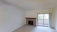 This two-floor space offers a functional and comfortable layout with notable design details throughout. The living area serves as a central feature of the home, highlighted by a classic brick fireplace with a wooden mantel that adds warmth and character to the room. Neutral-toned carpet covers the floor, complementing the clean white walls and creating a understated, versatile aesthetic. A textured popcorn ceiling adds a traditional finish overhead. A sliding glass door positioned adjacent to the fireplace floods the living space with natural light while providing direct access to a private balcony, where mature trees are visible in the background, offering a pleasant outdoor view and a sense of connection to nature. The overall ambiance of the living area is calm and inviting, with a straightforward design that provides a solid foundation for personalization. The two-floor layout suggests additional living or sleeping spaces on a separate level, allowing for a clear distinction between common areas and private quarters. The combination of the brick fireplace, natural light, and outdoor access makes this space both practical and welcoming.