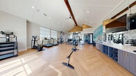 Located at 13000 Saginaw Blvd in Fort Worth, TX, this fitness center is a thoughtfully designed exercise space that combines modern aesthetics with a full suite of workout equipment. The room is anchored by dramatic vaulted ceilings with exposed wooden beams, creating an open and energizing atmosphere. Light-toned herringbone-patterned hardwood flooring runs throughout, adding a refined, contemporary touch to the space.

Natural light pours in through large windows along multiple walls, offering views of the surrounding outdoor greenery and keeping the environment bright and welcoming. Recessed and track lighting complement the natural light for well-lit workouts at any time of day.

The exercise floor is generously outfitted with a wide variety of equipment, including treadmills, rowing machines, a stair climber, an elliptical trainer, and stationary bikes for cardiovascular training. A multi-station cable and strength machine serves as the centerpiece for resistance training, accompanied by a dedicated free weights area with dumbbells and medicine balls neatly organized on racks.

A built-in storage and display unit along one wall houses additional accessories and is topped with a wall-mounted flat-screen television. A vibrant, flowing mural in soft aqua and cream tones adorns one wall, adding an artistic focal point that enhances the overall ambiance of this well-appointed fitness facility.