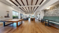 Located at 1501 Bellamy Pl in Louisville, KY, this single-floor amenity space offers a generously sized, open-concept layout designed for both socializing and relaxation. The interior is defined by soaring ceilings adorned with dark-stained exposed wood beams that contrast beautifully against crisp white walls, creating a warm yet modern aesthetic throughout.

Wide-plank hardwood flooring flows seamlessly across the entire space, unifying the distinct functional zones. A central kitchen island with a striking marble slab countertop serves as the focal point of the entertaining area, illuminated by elegant brass pendant lights. White cabinetry set within a dramatic arched stone alcove adds a refined architectural detail, while a cozy banquette nook with exposed brick walls provides a charming dining corner.

The lounge area features a sleek linear fireplace paired with a wall-mounted television and floor-to-ceiling black-framed windows that bathe the space in natural light and offer lush outdoor views. A billiards table anchors a dedicated recreation zone near the entrance, which opens through bold black French doors to an outdoor courtyard.

Additional seating areas and café-style tables are distributed throughout, making the space highly versatile. Recessed lighting, stone accent walls, and carefully curated décor complete this polished and inviting environment.