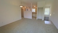 This single-floor residence offers a clean, open layout designed for comfortable everyday living. The main living area is finished with wall-to-wall carpeting in a neutral tone, creating a warm and cohesive foundation throughout the space. A decorative pendant light hangs above, adding a subtle focal point to the open room.

A low half-wall partition defines the transition between the living area and the kitchen without fully closing off the space, maintaining an airy, connected feel. The kitchen features white cabinetry along the walls, a dishwasher, and a window that invites natural light into the area, keeping the space bright and pleasant.

Ceiling-mounted flush lighting provides consistent illumination across the interior, complementing the natural light that filters in through multiple windows. A doorway visible from the main living area leads to additional rooms, suggesting a functional floor plan with clear circulation paths.

The overall ambiance is understated and practical, with light-toned walls and neutral finishes throughout that lend the space a clean, move-in ready character. The open sightlines between the living and kitchen areas make the floor plan feel spacious and well-connected, suitable for a variety of furnishing arrangements and lifestyles.