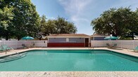 Located at 7684 Twin Beech Rd in Fairhope, Alabama, this single-story outdoor pool facility offers a well-maintained communal recreational space designed for relaxation and leisure. The centerpiece is a generously sized rectangular swimming pool with clear turquoise water, concrete pool decking, and entry via stainless steel handrails. Depth markers are visible along the pool edge, indicating a shallow-entry design suitable for a wide range of swimmers.

The facility features two distinct structural sections flanking the pool. On one side, a brick-accented building with a hip roof serves as a utility or changing room structure, finished with blue-gray siding above a red brick base. Wall-mounted light fixtures provide functional outdoor lighting. On the opposite side, a covered pavilion with white columned posts and a coordinating hip roof offers a shaded gathering area, complete with French doors opening to an interior space — ideal for events or community use.

The entire pool area is enclosed by a white privacy fence, creating a secure and cohesive environment. Lounge chairs and teal patio umbrellas are positioned around the deck, enhancing the resort-like atmosphere. Mature deciduous trees surround the perimeter, providing natural shade and a lush, serene backdrop. The overall setting reflects a well-appointed community amenity in the heart of Fairhope.