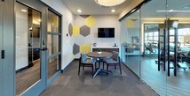 This single-floor amenity space combines functionality and modern design in a cohesive, well-appointed layout. The interior is immediately distinguished by its bold hexagonal wall graphics in yellow, gray, and charcoal tones, which carry consistently throughout the space and lend it a vibrant, contemporary character.

The lounge and gathering area serves as a central hub, furnished with a round wooden dining table and cushioned chairs beneath a large circular pendant light. A wall-mounted flat-screen TV and a low credenza with decorative accents complete the area, making it well-suited for casual meetings or relaxation. Recessed lighting and a neutral carpet tile floor contribute to the warm, inviting ambiance.

Adjacent to the lounge, a dedicated business center station offers a desktop computer and printer along a clean, built-in countertop with bar-height seating — a practical workspace for productivity needs.

A striking full-height glass wall partition with a frosted linear pattern separates the lounge and business area from the fitness center, maintaining visual openness while defining each zone. The fitness center is equipped with a row of treadmills, ellipticals, and strength training equipment, and benefits from large windows that allow natural light to fill the space.

Blue-framed glass entry doors with transom windows provide a polished, welcoming entrance to the entire amenity suite.