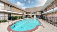 Located at 8900 Chimney Rock Rd in Houston, Texas, this outdoor amenity space centers around a generously sized swimming pool set within a well-maintained courtyard. The pool is framed by a classic brick-trimmed surround and is enclosed by ornamental wrought iron fencing, giving the area a defined and secure atmosphere. Teal lounge chairs and patio furniture are thoughtfully arranged poolside, offering ample space for relaxation and socializing.

The courtyard is enclosed on multiple sides by a two-story building featuring warm brick exterior walls, covered walkways with white-trimmed overhangs, and individual unit balconies with decorative iron railings. Arched brick detailing adds a classic architectural character to the ground-floor passages. Manicured shrubbery lines portions of the perimeter, contributing a touch of greenery that softens the hardscape.

A dedicated pool house structure anchors one end of the courtyard, displaying posted pool rules and providing access to pool amenities. An additional shallow wading area is visible near the pool house entrance. Mature trees within the courtyard provide natural shade, while the open sky above allows for abundant natural light. The overall ambiance is inviting and well-organized, making this courtyard pool area a notable communal feature of the property.