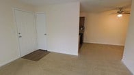 This single-floor residential unit offers a straightforward, functional layout designed for comfortable everyday living. The main living area is finished with plush wall-to-wall carpeting in a neutral beige tone, creating a warm and inviting atmosphere. A partial wall divides the living space from the kitchen area, providing a sense of separation while maintaining an open, connected feel throughout the floor plan. The entryway transitions seamlessly from wood-style laminate flooring into the carpeted living space, adding a practical and visually distinct threshold. A white paneled door near the entry provides access to a closet, offering convenient storage. The kitchen, partially visible from the living area, features dark wood cabinetry, a tile backsplash with a subtle pattern, and dark appliances, lending a classic and cohesive look to the cooking space. The walls throughout are painted in a clean, light neutral tone, contributing to a bright and airy ambiance. The textured ceiling adds a subtle architectural detail consistent across the space. Overall, the unit presents a clean, move-in ready environment with a practical layout suited for a range of living arrangements.