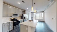 Located at 4206 Simcoe Ln in Louisville, Kentucky, this single-floor residence offers a thoughtfully designed open-concept layout with contemporary finishes throughout. The kitchen serves as the heart of the home, featuring gray shaker-style cabinetry, a white quartz island with an undermount sink, subway tile backsplash, a built-in wine rack, and a full suite of stainless steel appliances including a French door refrigerator, electric range, and microwave. Elegant pendant lighting hangs above the island, complementing the recessed lighting overhead. Dark hardwood-style flooring flows seamlessly through the main living areas, connecting the kitchen to a generous living space with crown molding accents and a ceiling fan. Large windows and a glass door open to a private balcony, offering views of the surrounding neighborhood and abundant natural light. The bedroom features plush carpeting, a wide window with blinds, and access to a spacious walk-in closet with wire shelving. The bathrooms are well-appointed, with one showcasing a glass-enclosed shower adorned in large-format marble-look tile with brushed nickel fixtures, and another featuring a long vanity with a white countertop and a wide framed mirror. An in-unit washer and dryer area adds everyday convenience.