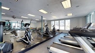 This fitness center is designed to support a comprehensive workout experience within a single, open-plan floor. The space is outfitted with a variety of cardio equipment, including treadmills, elliptical trainers, and a stationary bike, arranged to allow comfortable movement between machines. A dedicated free weights area lines one wall, featuring a full rack of color-coded dumbbells suited for a range of strength training exercises. Full-length mirrors span an entire wall, enhancing the sense of space and allowing users to monitor their form during workouts. The flooring consists of durable, dark rubberized material designed to absorb impact and support heavy equipment. Overhead, recessed flat-panel lighting fixtures provide bright, even illumination throughout the room, complemented by natural light streaming in through a generous bank of windows along one wall. These windows also offer views of the surrounding exterior, adding an open, airy quality to the environment. The textured ceiling and neutral wall tones contribute to a clean, functional aesthetic. Overall, the fitness center balances practical utility with a welcoming atmosphere, making it a well-rounded facility for daily exercise.