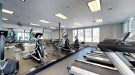 This fitness center is designed to support a comprehensive workout experience within a single, open-plan floor. The space is outfitted with a variety of cardio equipment, including treadmills, elliptical trainers, and a stationary bike, arranged to allow comfortable movement between machines. A dedicated free weights area lines one wall, featuring a full rack of color-coded dumbbells suited for a range of strength training exercises. Full-length mirrors span an entire wall, enhancing the sense of space and allowing users to monitor their form during workouts. The flooring consists of durable, dark rubberized material designed to absorb impact and support heavy equipment. Overhead, recessed flat-panel lighting fixtures provide bright, even illumination throughout the room, complemented by natural light streaming in through a generous bank of windows along one wall. These windows also offer views of the surrounding exterior, adding an open, airy quality to the environment. The textured ceiling and neutral wall tones contribute to a clean, functional aesthetic. Overall, the fitness center balances practical utility with a welcoming atmosphere, making it a well-rounded facility for daily exercise.