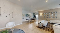 This single-floor space offers a generously sized, open-plan layout that seamlessly integrates a living area and kitchen into one cohesive environment. The interior is finished with large-format neutral tile flooring throughout, contributing to a clean, contemporary aesthetic that flows across the entire space.

The living area is expansive and thoughtfully arranged, featuring plush white leather sofas centered on a patterned area rug, a wall-mounted flat-screen television, and a striking grid of framed mirror-style wall art that adds a decorative focal point. A pool table with a chrome frame anchors a secondary activity zone, reinforcing the social and entertainment-oriented character of the space. Recessed lighting and wall sconces provide layered illumination throughout.

The kitchen is well-appointed with rich dark wood cabinetry, granite countertops, stainless steel appliances, and a curved peninsula with seating for multiple guests. A ceiling fan overhead and natural light from adjacent windows create a bright, airy atmosphere in this area.

A welcoming entry foyer with glass double doors, a statement pendant light fixture, and views to the exterior completes the space, providing an impressive first impression upon arrival.