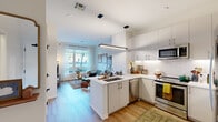 This thoughtfully designed single-floor residence offers a seamless blend of modern aesthetics and functional living. The open-concept kitchen and living area serves as the heart of the space, where crisp white cabinetry, a classic subway tile backsplash, and stainless steel appliances define a well-equipped kitchen anchored by a central island with an undermount sink. A linear pendant light above the island adds a contemporary touch, while light hardwood flooring flows consistently throughout, unifying the space. The living area benefits from generous natural light through large windows dressed with sheer curtains, creating a bright and welcoming atmosphere. Two bedrooms each feature oversized windows that frame outdoor views and allow abundant daylight to fill the rooms, complemented by clean white walls that lend a calm, neutral backdrop. A flexible secondary room functions well as a home office or sitting area, complete with elevated ceiling height and warm, diffused lighting. The bathroom is finished with floor-to-ceiling white subway tile and a glass-enclosed walk-in shower, offering a clean and modern design. Throughout the residence, the consistent use of light tones, hardwood flooring, and contemporary fixtures creates a cohesive and inviting living environment.