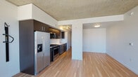 This single-floor unit showcases a contemporary industrial-modern aesthetic defined by its signature exposed concrete ceiling, which contrasts beautifully with clean white walls and warm light hardwood flooring that extends throughout the entire space. The open layout connects the kitchen and living areas seamlessly, creating a cohesive and functional flow.

The kitchen is well-appointed with dark espresso cabinetry, stainless steel appliances including a French door refrigerator with an ice and water dispenser, a gas range, built-in microwave, and a single-basin sink. Track lighting illuminates the galley-style workspace efficiently.

The living area is bright and open, benefiting from expansive floor-to-ceiling windows that frame impressive panoramic city views, filling the space with abundant natural light. A wall-mounted HVAC unit sits beneath the windows, integrated neatly into the design.

Additional spaces include a generously sized secondary room, suitable as a bedroom or flexible living space, featuring the same hardwood floors and concrete ceiling. The bathroom is clean and minimalist, with white fixtures and hardwood flooring. A stacked washer and dryer unit is also included within the unit, as visible in the 3D overview, adding practical convenience to this modern urban residence.