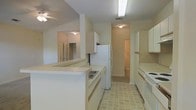 This single-story space centers around a functional kitchen that serves as the heart of the layout. The kitchen is designed with ample white cabinetry lining both sides of the galley-style corridor, providing generous storage throughout. A peninsula counter extends into the adjacent living area, creating a natural divide between spaces while offering additional workspace and casual seating potential. White appliances are included, featuring a refrigerator, range, and dishwasher, all integrated seamlessly into the cabinetry layout. The flooring in the kitchen features neutral tile in a classic pattern, offering durability and ease of maintenance. Overhead, a flush-mounted light fixture ensures consistent illumination throughout the kitchen corridor. A textured ceiling adds subtle visual interest throughout the space. The layout connects openly to a living area, where a ceiling fan enhances airflow and comfort. A mirrored closet is visible from the living space, contributing to a sense of depth and brightness. Doorways lead to additional rooms beyond the kitchen, suggesting a practical and well-connected floor plan. The overall palette of whites and soft neutrals lends a clean, cohesive ambiance throughout the space.