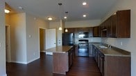 This single-story space showcases an open-concept kitchen designed for both functionality and style. The kitchen is anchored by a central island with a laminate countertop, offering additional workspace and casual seating potential. Dark espresso-toned cabinetry lines the walls, providing ample storage, and is complemented by matching lower cabinets that extend along two walls in an L-shaped configuration. Stainless steel appliances, including a refrigerator, electric range, and over-the-range microwave, lend a modern touch to the space. A double-basin sink is situated along the far counter run, positioned beneath upper cabinetry. Recessed lighting is installed throughout the ceiling, supplemented by two elegant pendant lights that hang above the island, adding warmth and focused illumination. Rich, dark hardwood flooring runs throughout the space, creating a cohesive and polished aesthetic. A doorway visible in the background hints at additional rooms beyond, while a closet with bi-fold doors is partially visible, suggesting nearby storage or utility space. The overall ambiance is contemporary and well-appointed.