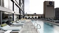 This rooftop pool deck delivers a refined outdoor experience elevated above the city skyline. The centerpiece is a generously sized rectangular swimming pool with crystal-clear water, framed by large-format light grey stone pavers that extend across the entire deck. Rows of contemporary cushioned chaise lounges line both sides of the pool, providing ample space for relaxation and sunbathing. The lounge chairs feature clean white frames paired with soft teal and grey cushions, accented by accent chairs in warm yellow tones that add a pop of color to the palette. Potted trees and planters are thoughtfully placed throughout the deck, introducing greenery that softens the urban setting. The deck is bordered by open railings that preserve unobstructed views of the surrounding cityscape, including notable skyline elements visible in the distance. A modern multi-story building with floor-to-ceiling glass windows flanks one side of the deck, reflecting the contemporary architectural style of the property. The overall ambiance is sleek, sophisticated, and resort-inspired, making this rooftop deck an ideal setting for both leisurely lounging and social gatherings in an urban environment.