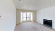 This single-floor space offers a clean, functional layout with a warm and neutral aesthetic throughout. The main living area is a standout feature, characterized by plush wall-to-wall carpeting in a soft, neutral tone that adds comfort underfoot. A prominent bay window serves as the focal point of the room, allowing an abundance of natural light to flood the interior and offering outward views of the surrounding greenery. The multi-panel bay window design creates a subtle architectural interest, adding dimension to the otherwise clean-lined walls. The ceiling features a classic textured finish, adding depth and visual character to the space. Walls are finished in a crisp, light neutral tone, creating a versatile backdrop that complements a variety of furnishing styles. Tile flooring is visible at the room's edge, suggesting a transition to an adjacent area with hard surface flooring. HVAC venting is integrated into the ceiling, ensuring functional climate control throughout. Overall, the space presents a bright, airy ambiance with practical finishes and a welcoming feel suited to a range of uses.