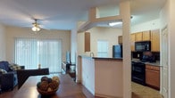 Located at 7601 Gateway Blvd in Live Oak, Texas, this single-story unit offers a well-designed open-concept floor plan that seamlessly connects the main living areas. Rich hardwood-style flooring flows throughout the living, dining, and kitchen spaces, creating a cohesive and inviting atmosphere.

The living room is bright and comfortable, featuring a ceiling fan with light fixture and a large sliding glass door dressed with vertical blinds that allows natural light to fill the space. The dining area is positioned adjacent to the living room, providing a natural transition between the two spaces.

The kitchen is well-equipped with warm wood cabinetry, dark granite-style countertops, and black appliances including a refrigerator, electric range with microwave, and dishwasher. A pass-through architectural detail connects the kitchen to the dining area, adding an open, airy feel. The kitchen also benefits from a window that brings in additional natural light.

The bedroom is generously sized with plush carpeting, a ceiling fan, and two separate closets offering ample storage. The bathroom is spacious and functional, featuring rich wood-toned cabinetry, a dual-sink vanity with granite-style countertops, and hardwood-style flooring that extends throughout the space. Overall, the unit presents a practical and comfortable living environment.