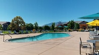 This outdoor amenity area centers around a generously sized freeform swimming pool with clear turquoise water, accessible via stainless steel entry rails on multiple sides. The pool's curved design creates an inviting focal point within the space, surrounded by a broad, stamped concrete sun deck that provides ample room for relaxation and recreation.

The deck is well-furnished with reclining lounge chairs positioned to take full advantage of the abundant natural sunlight, while vibrant umbrella-shaded seating clusters offer comfortable spots for those seeking shade. Decorative planters featuring seasonal flowers and mature ornamental trees are thoughtfully integrated throughout the landscape, adding color and texture to the environment.

A covered pavilion or clubhouse structure is situated at the edge of the pool area, providing a sheltered gathering space that complements the outdoor setting. The entire area is enclosed by a black metal fence, ensuring both security and a clean visual boundary, while still allowing unobstructed views of the surrounding mountain range and blue skies beyond.

Lush lawn areas and carefully maintained shrubs and trees border the perimeter, enhancing the natural ambiance. The overall design balances functionality with aesthetics, making this a well-rounded outdoor retreat suited for both active use and leisurely enjoyment in a scenic, open-air setting.